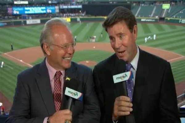 two guys in suits broadcasting in pres box in front of baseball field