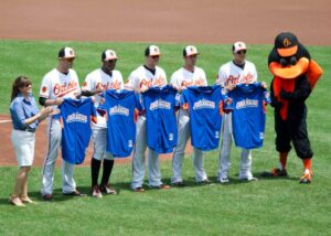 orioles players standing in line on field holding jerseys with mascot and woman clapping